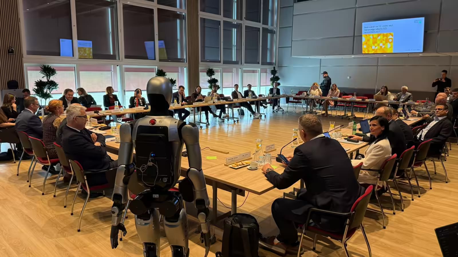 Humanoidni robot MAX stoji ob mizi na seji upravnega odbora Gospodarske zbornice Slovenije, udeleženci ga opazujejo