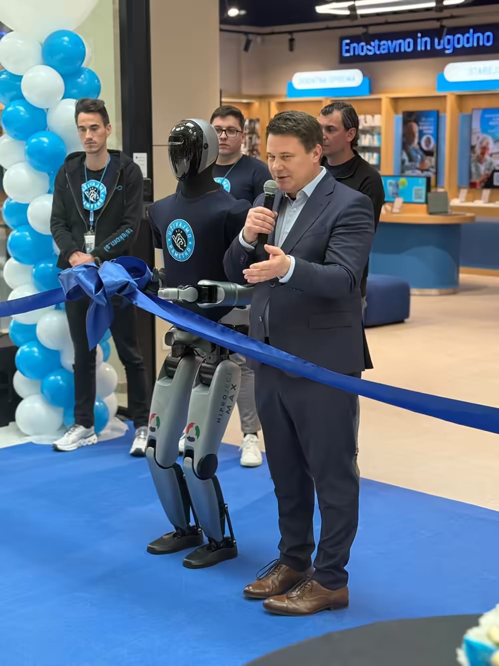 Humanoidni robot MAX ob modrem traku na odprtju poslovalnice, poleg njega govornik z mikrofonom