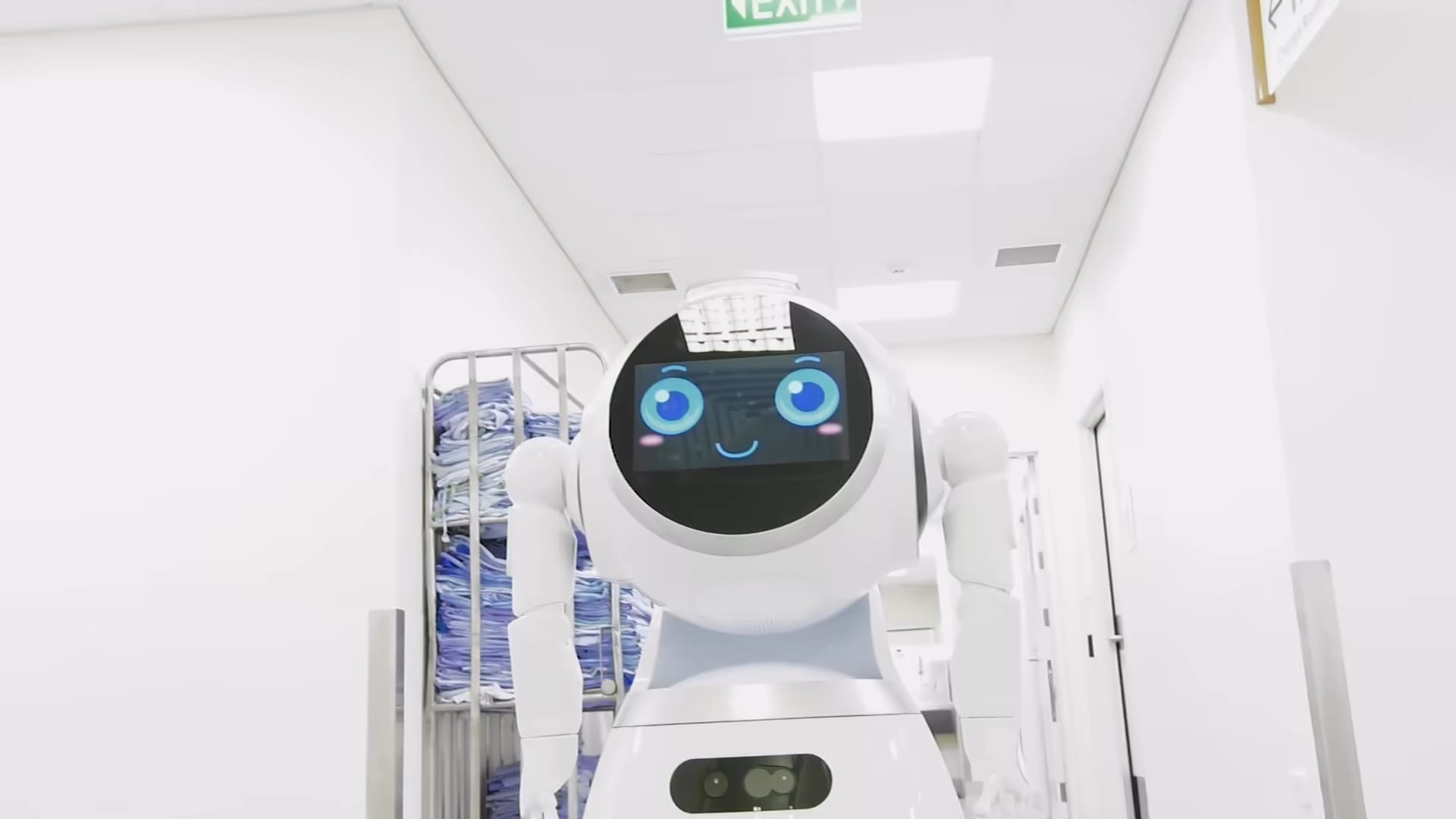 Humanoidni robot z zaslonom namesto obraza se pelje po bolniškem hodniku mimo vozičkov s perilom.