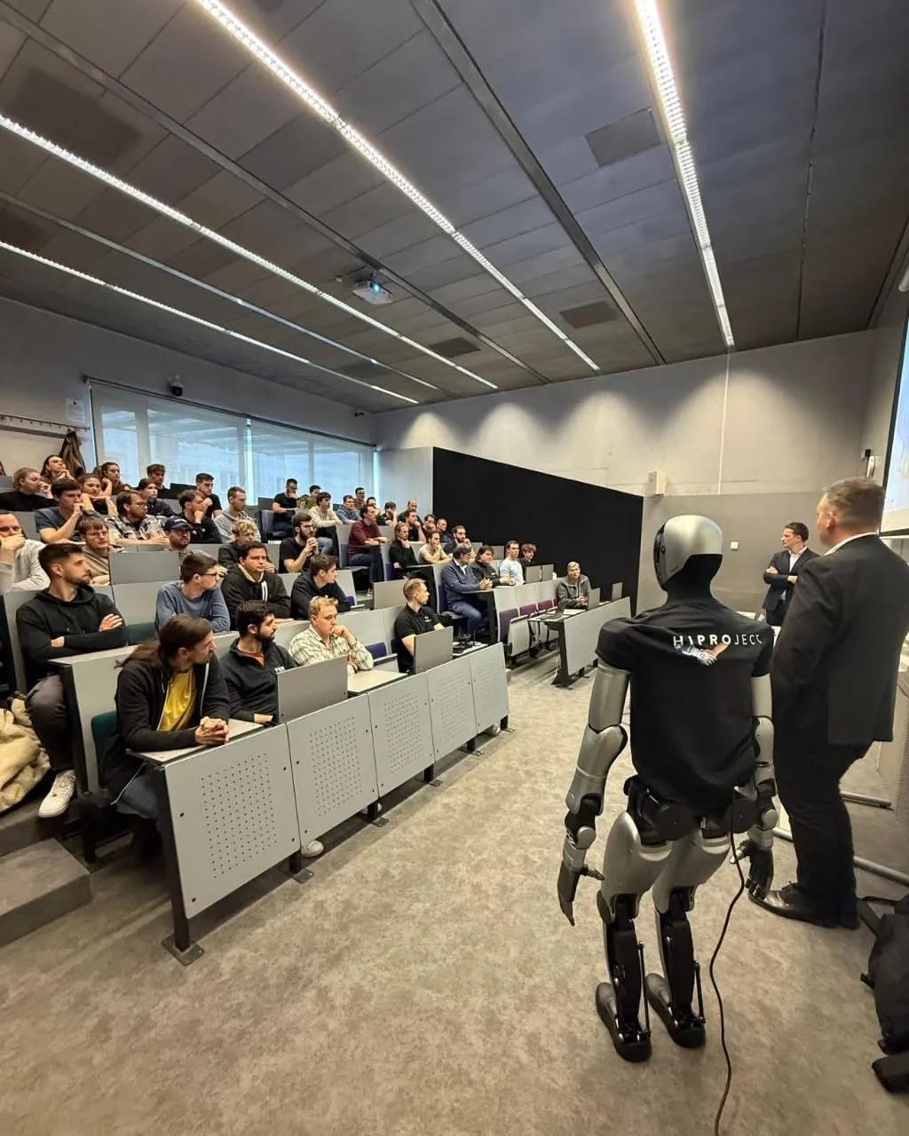 Humanoidni robot v majici HIPROJECT pred polno predavalnico študentov