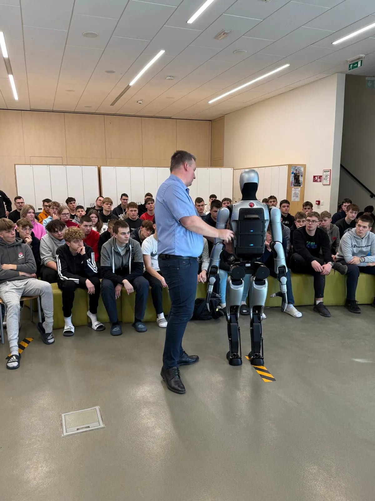 Predavatelj ob humanoidnem robotu pred dijaki v šolski avli