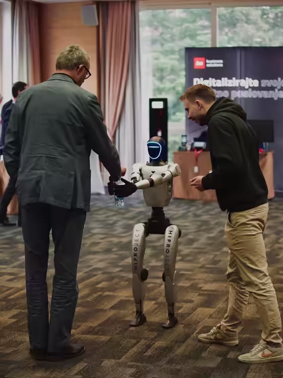 Humanoidni robot NEO med dvema obiskovalcema na poslovnem dogodku, robotu izroča stekleničko
