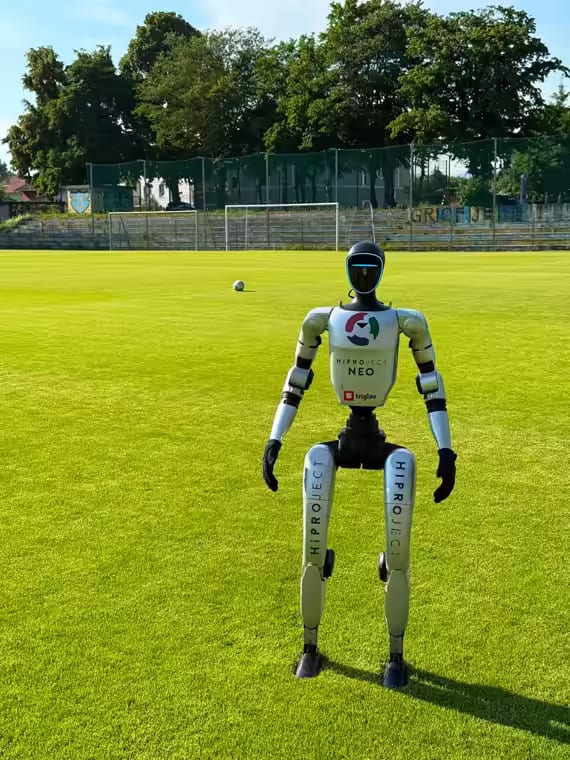 Humanoidni robot NEO stoji na nogometnem igrišču, v ozadju gol in žoga