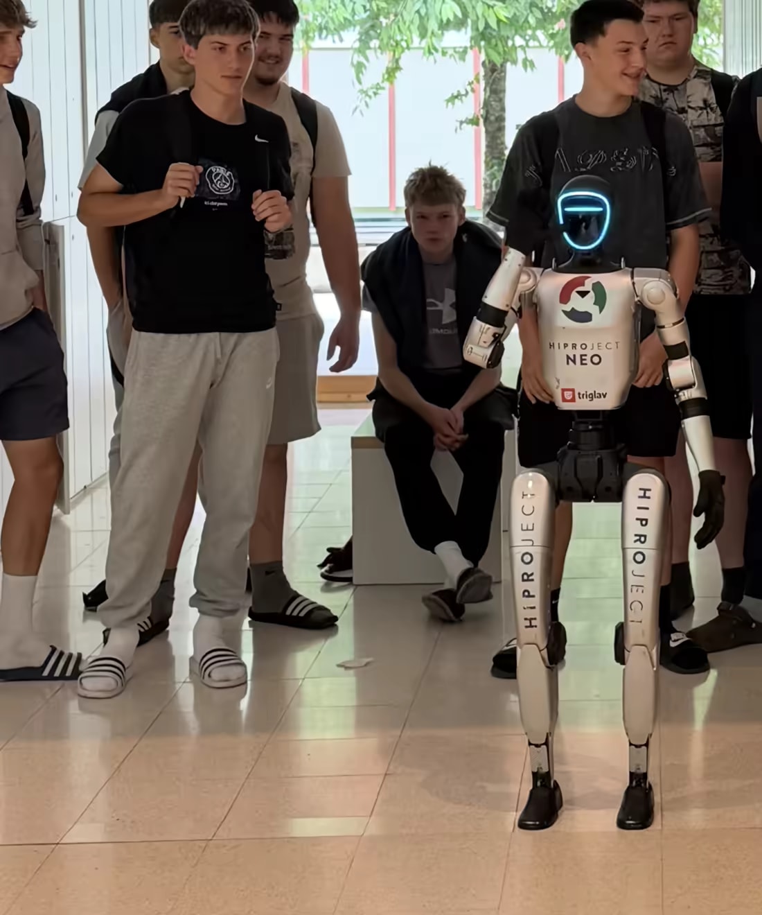 Robot NEO stoji med skupino dijakov na šolskem hodniku.