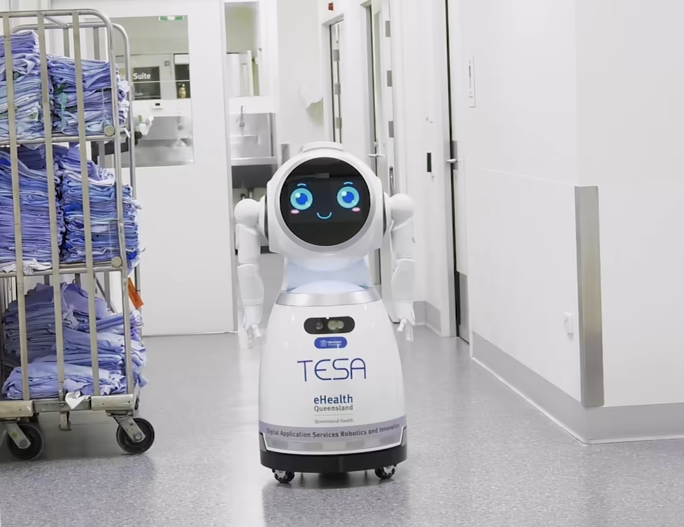 Robot CRUZR na bolniškem hodniku, za njim voziček s perilom.