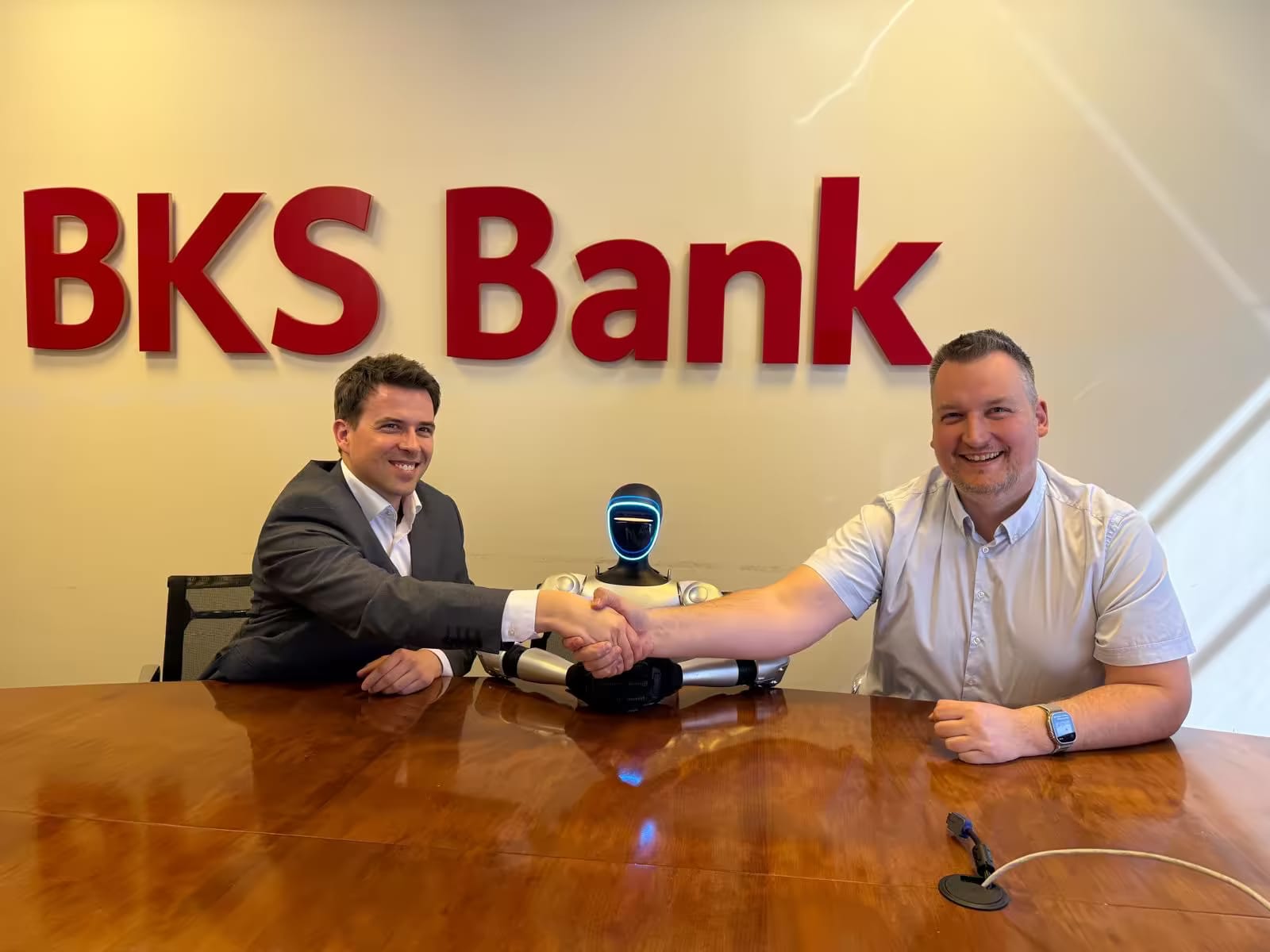 Stisk rok ob sklenitvi sodelovanja v prostorih BKS Bank, med sogovornikoma sedi humanoidni robot.